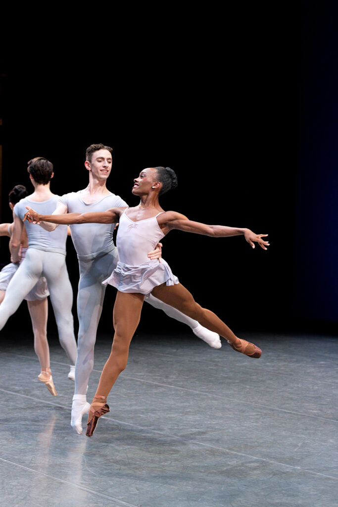 A Black ballet dancer wearing a skirted leotard and pointe shoes is partnered by a white dancer. Both are jumping in arabesque.