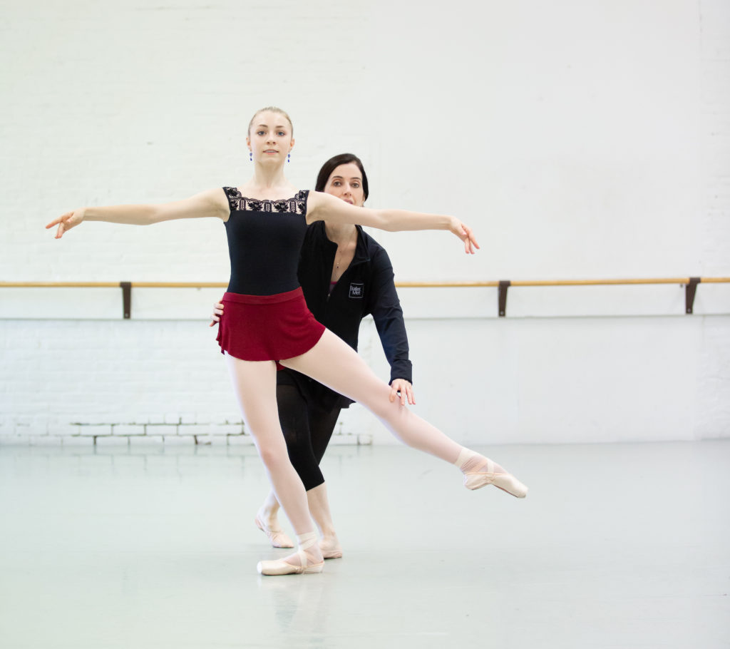 A teen female ballet dancer brushes her left leg to second while her teacher standing behind her corrects her alignment.