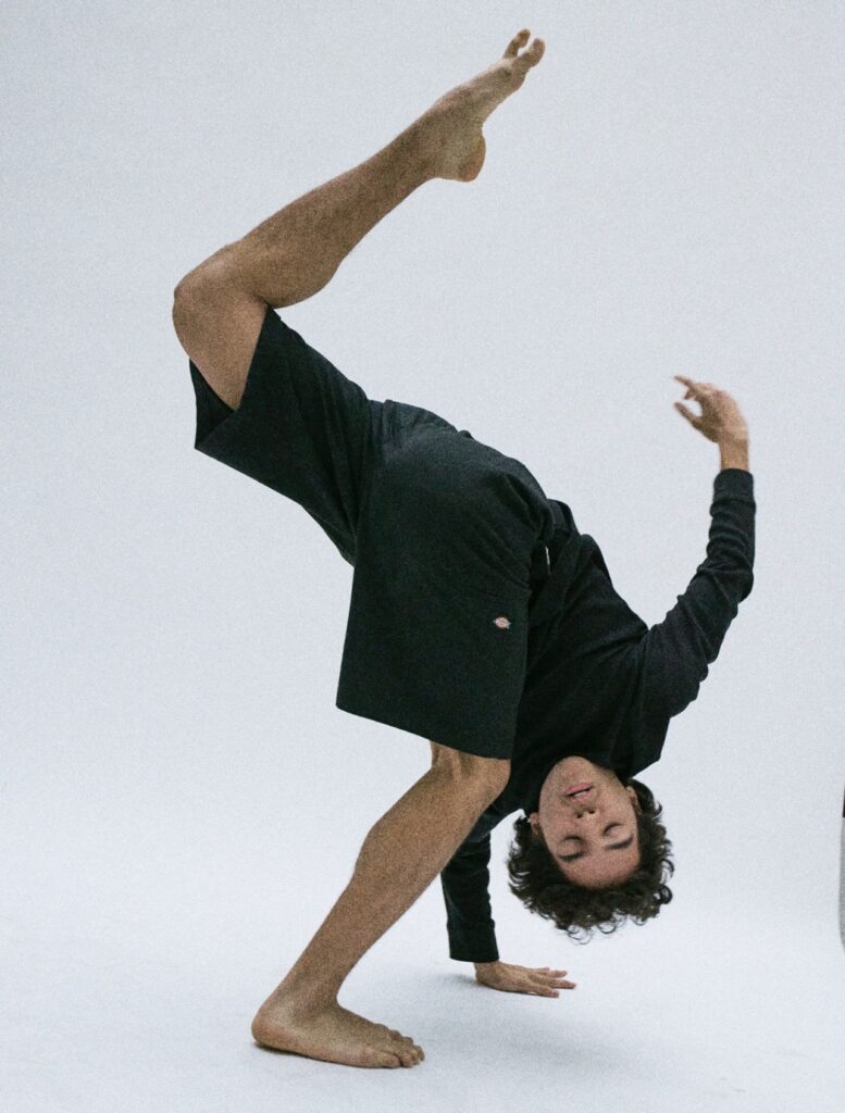 Alejandro Vargas posing against a white backdrop. He bends forward with one leg lifted in the air in attitude