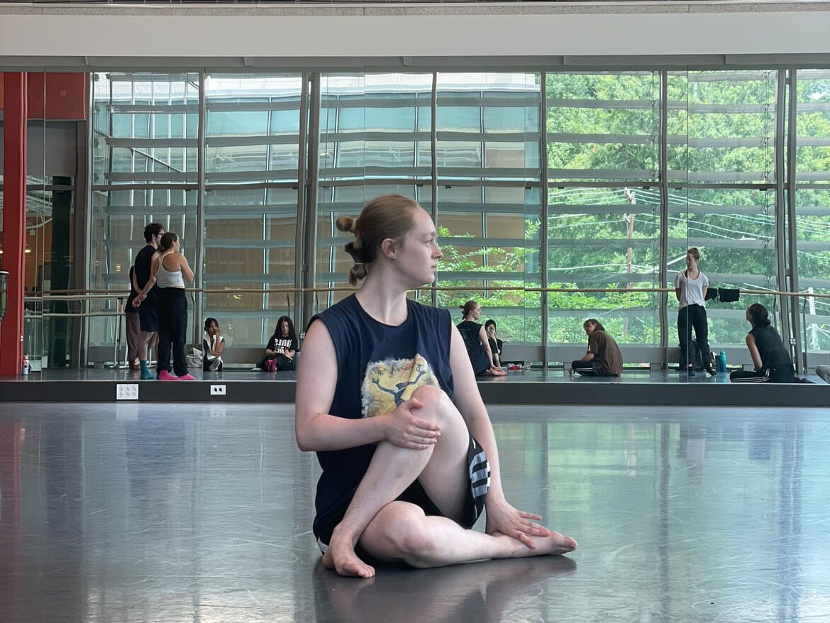 Aubrey Mailloux sitting on the floor of a studio stretching with one leg crossed over the other