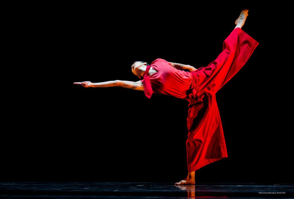 A female dancer wearing a red costume reaches laterally with one arm while kicking the opposite leg