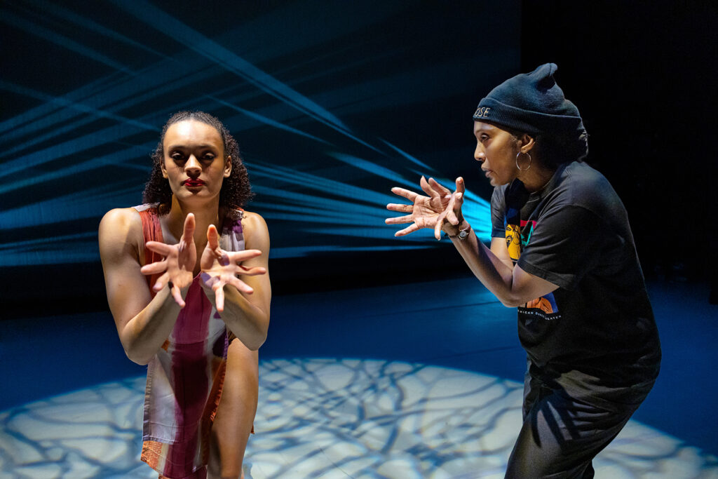 A Black dancer extends her hands toward the camera, fingers splayed. To her left, a Black instructor demonstrates the same move, coaching the performer.