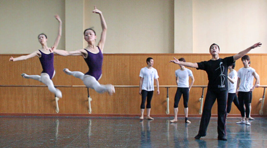 A man in a black t-shirt and pants leads a ballet class. Two female dancers in black leotards and pink tights leap through the studio.