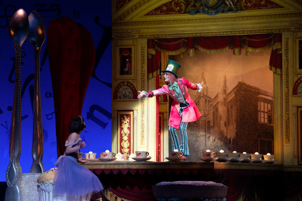 On a highly-decorated stage, the Mad Hatter reaches out towards Alice, who wears a purple dress.