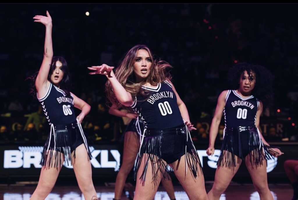 On a basketball court, a group of dancers perform high-energy choreography together in costumes that look like basketball uniforms. Kayla Cruz, in the center, flicks her and and pops her hip, her hair flying.