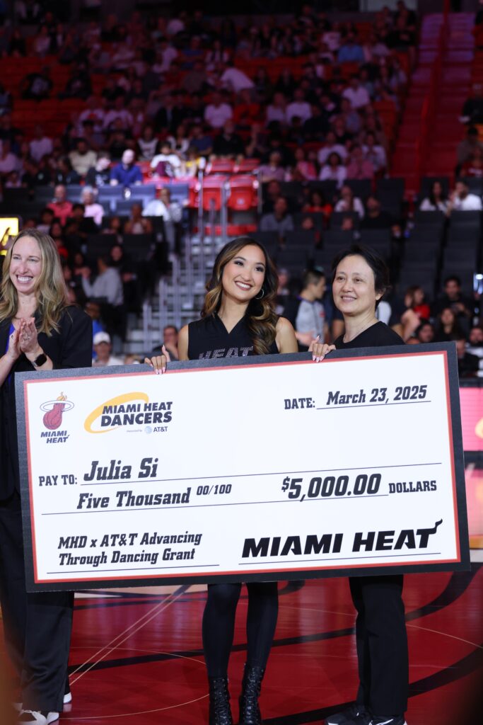 In a crowded basketball stadium, Julia Si smiles and holds a large cardboard check as two women on either wide of her smile and celebrate.