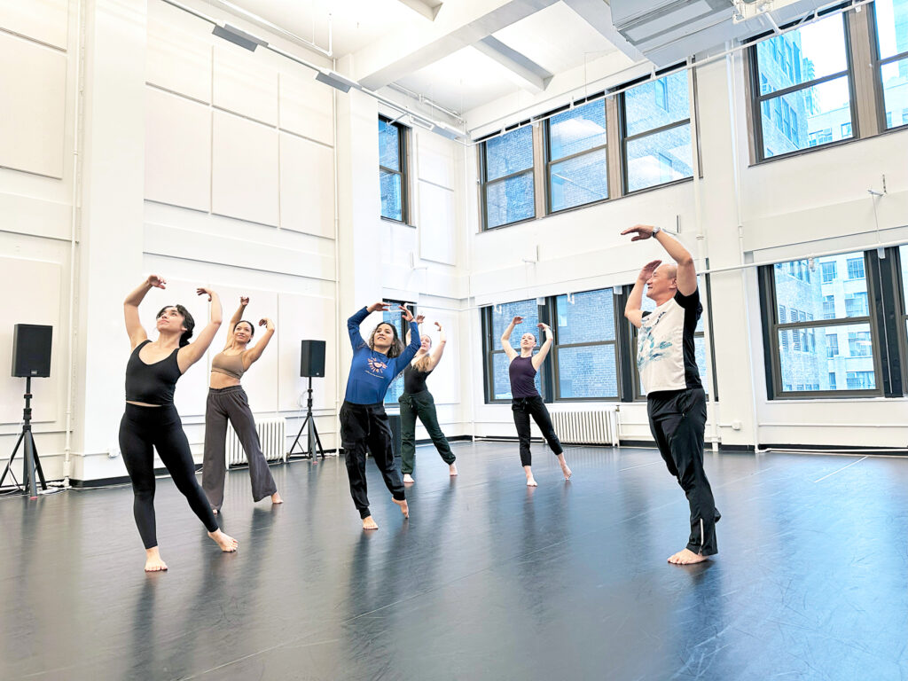 An Asian man in black pants and a white t-shirt demonstrates a dance movement with back arched and arms above the head. A group of dancers mimics his pose.