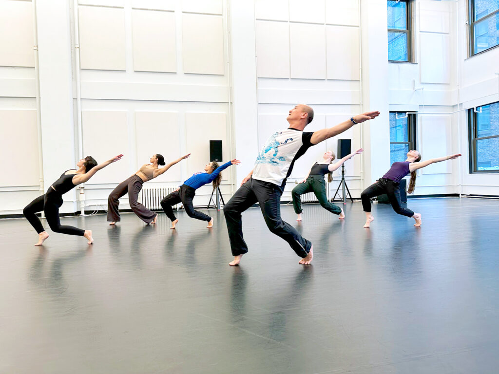 An Asian man wearing black pants and a white t-shirt leans back into a bent-kneed hinge position, one arm extended back. A group of dancers is behind him in the same pose.