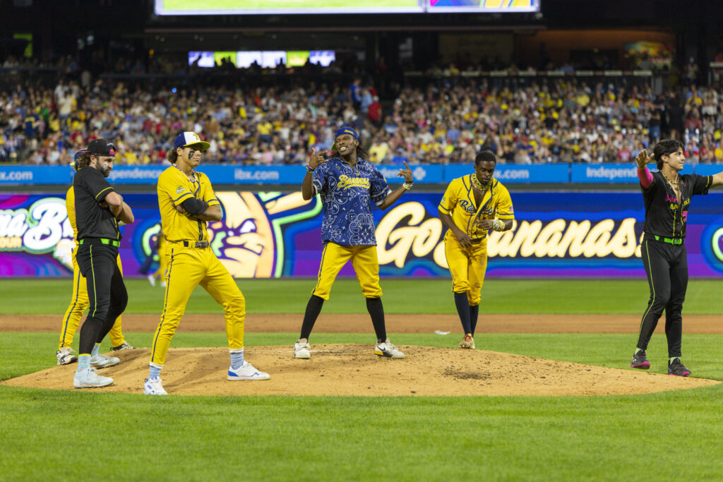 A group of Savannah Bananas baseball players run into place or pose with arms crossed. Maceo Harrison, in the middle, speaks to the crowded stadium and gestures with his hands.