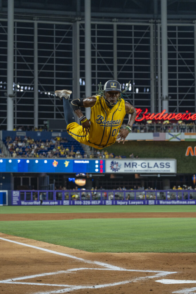 On a baseball field, Maceo Harrison flies through the air, body parallel with the field, as he puts his hands on his hips and bends his legs up toward the sky. He wears a bright yellow Savannah Bananas uniform.