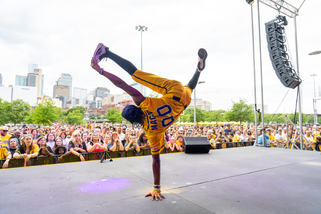 Maceo Harrison breaks onstage, using one hand to support his weight as he kicks up and over to touch his other hand with one foot, the other leg suspended up in the air. 