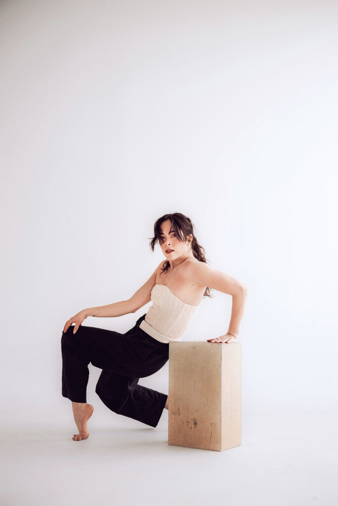 A white dancer with brown hair wearing black pants and a white top poses in a low crouch next to a tan box, against a white backdrop. 