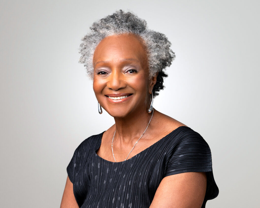 A professional headshot of a smiling older Black woman with gray hair, wearing a black top against a white background.