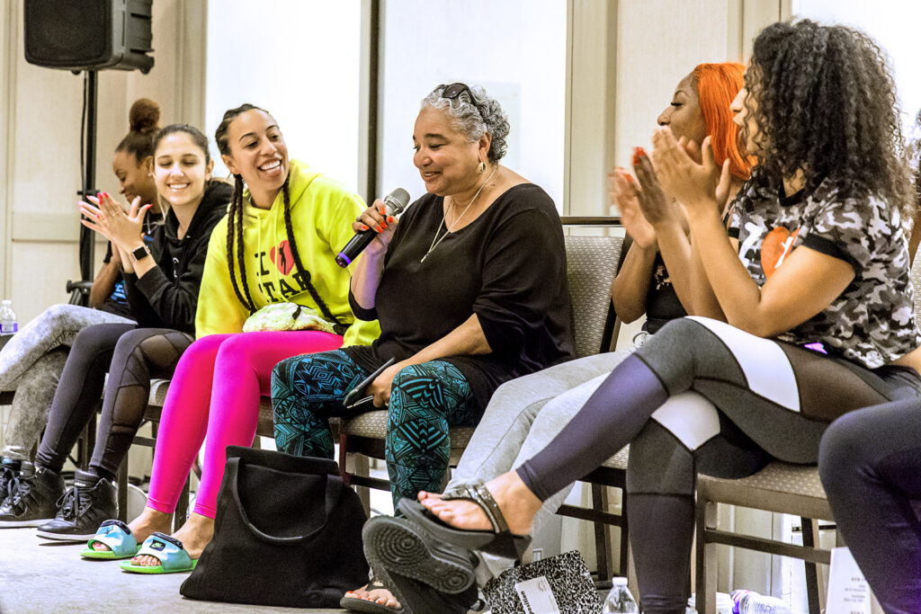 A group of women sits in a row with their backs to a dance studio mirror, having a spirited discussion. The Black woman in the center of the row holds a microphone.