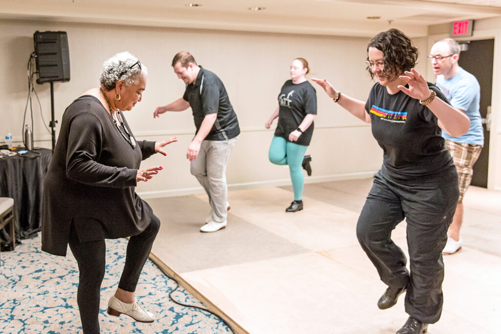 A Black woman with white hair wearing all black teaches four adult tap dancers a step. All stand on one leg with the other foot lifted off the floor.