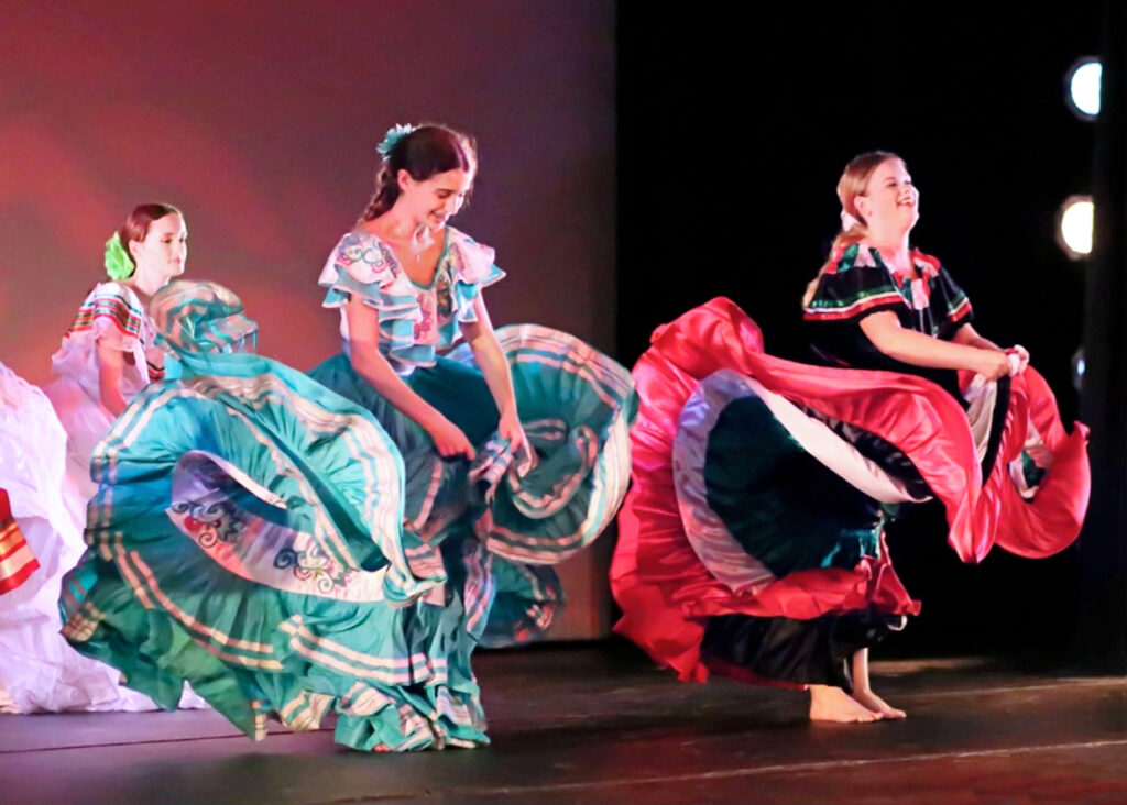 Teenaged dancers wear colorful cultural costumes that include large, ruffled skirts.