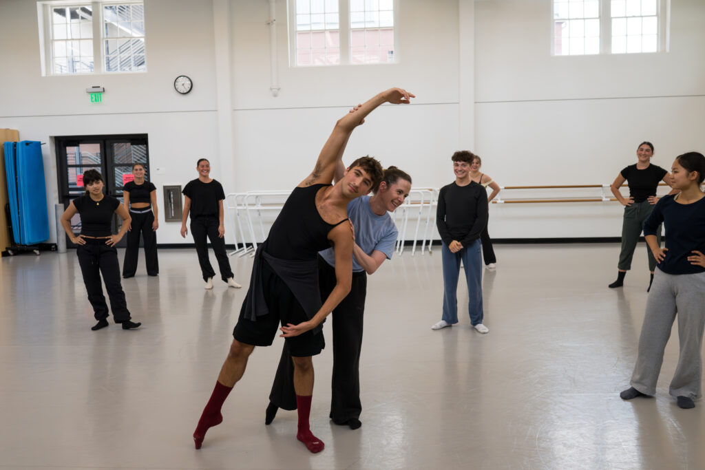 A female instructor guiding a student as performs a tendu écarté. Other dancers look on in the studio.