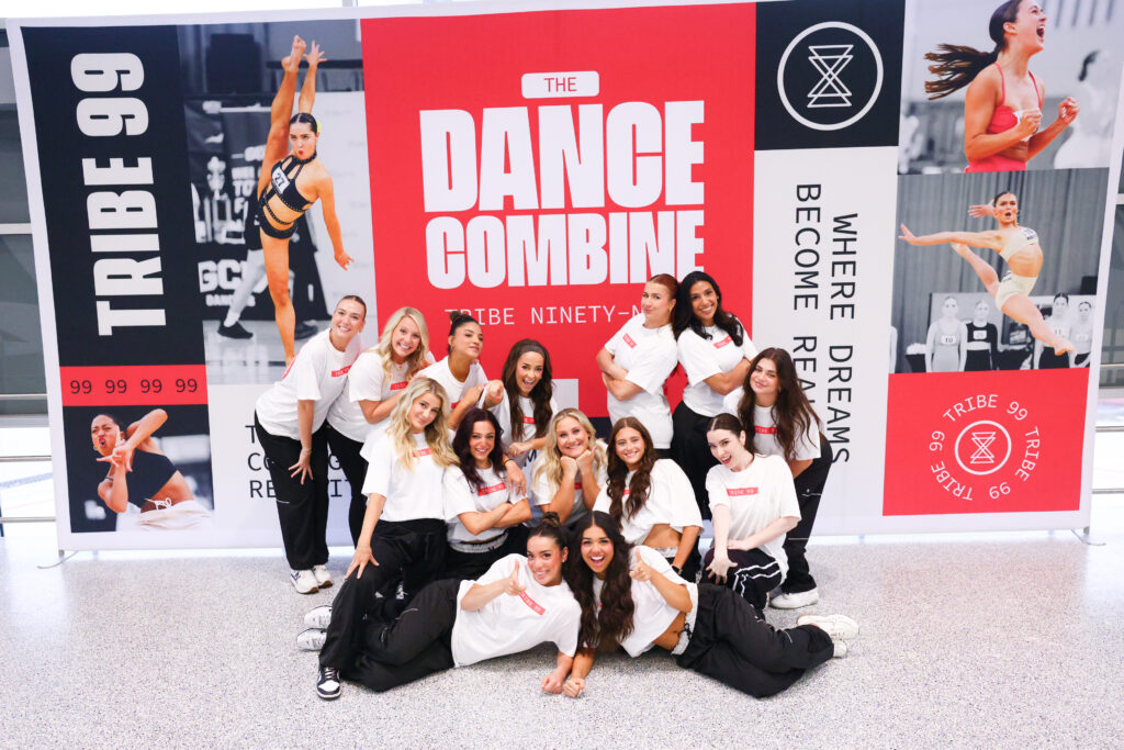 In front of a large Tribe 99 poster, a group of female dance staff pose for a group photo in matching t-shirts and black pants.