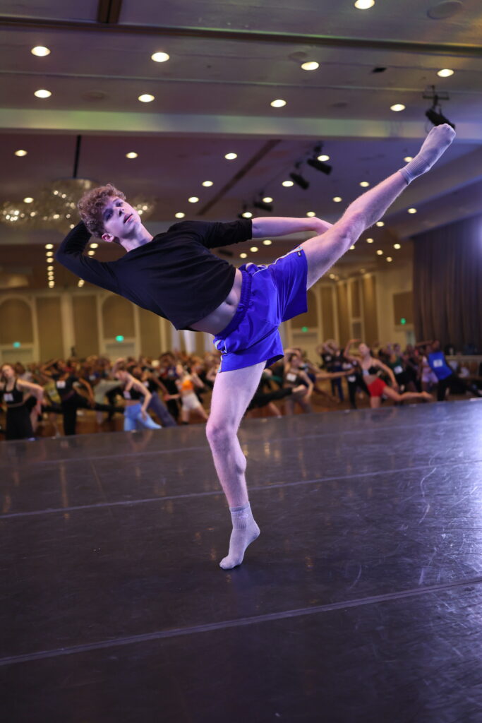 Cameron Jansen balances on forced arch in a tilted arabesque line, his lifted leg facing forward and directly to the side. He reaches one arm back behind his leg and the other bends up behind his head as he leans his upper body over past his bent supporting leg. He dances onstage in front of a large group of dancers at a convention in a ballroom.