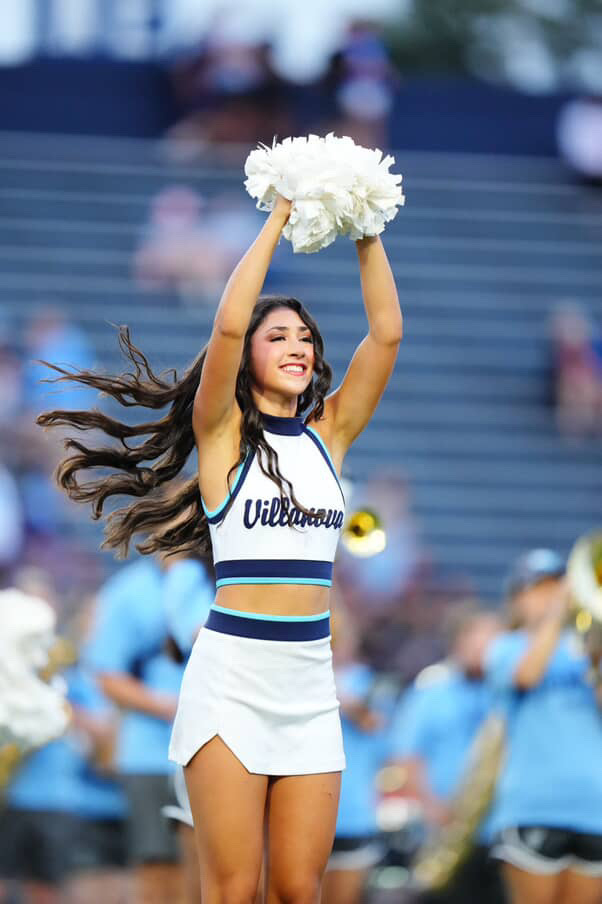 A female dancer wearing a blue and white cheer uniform. She smiles and waves poms overhead.