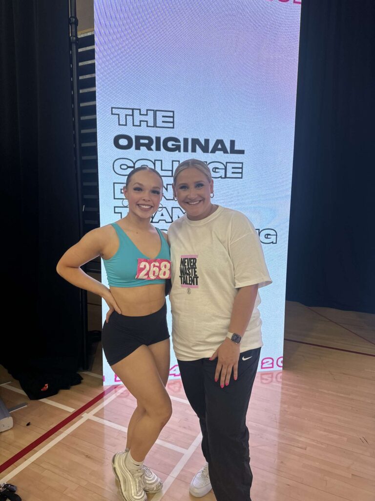 Rayna Reid and Carson Rowe pose for a photo at a DANCE COMBINE event. Reid wears dancewear and an audition number, and Rowe wears a staff t-shirt and black pants.