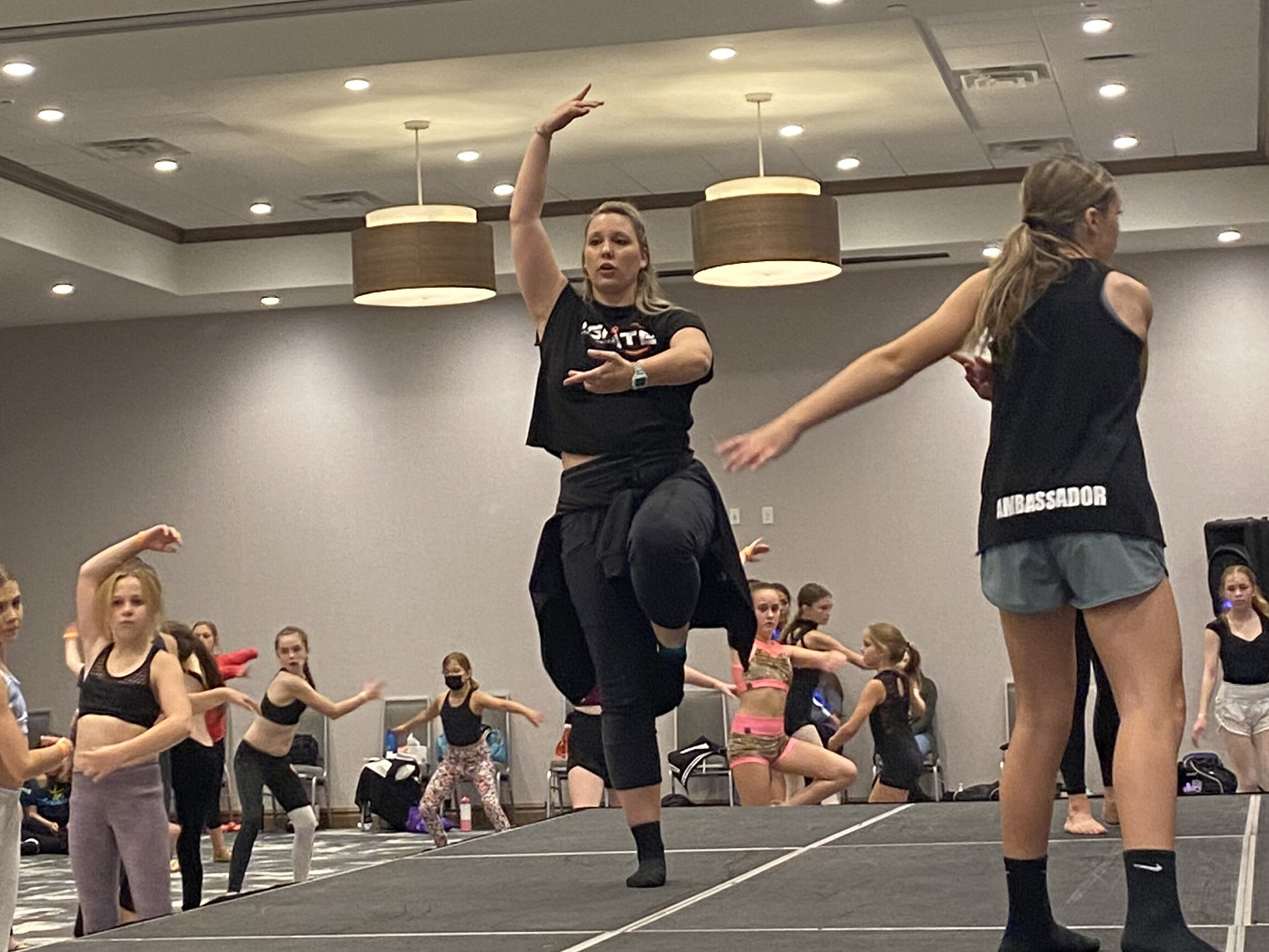 A female instructor demonstrating on stage. She lifts her left knee into a parallel passe, one arm in 5th position, and one arm in 1st position. 
