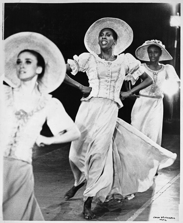 A black and white photo of three Black female dancers wearing white costumes that consist of broad-brimmed hats, blouses, and long skirts.
