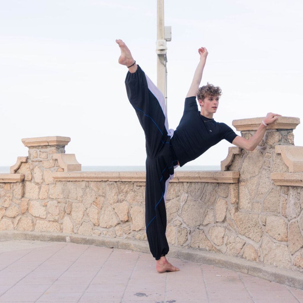 On an elevated outdoor patio, Cameron Jansen does a tilt with a slightly-bent top leg as he rests one hand on top of a stone wall. He reaches his other arm overhead and looks out into the distance.