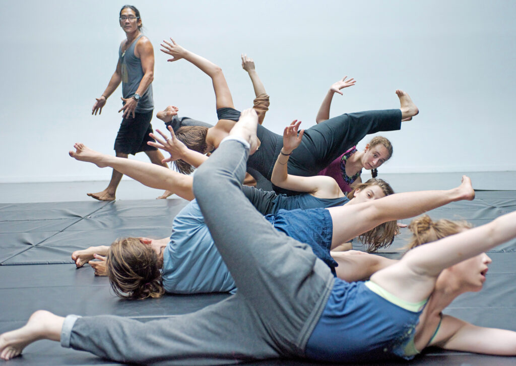 A group of dancers are on the floor, arms and legs outstretched. A man walks in the background, observing their movements.