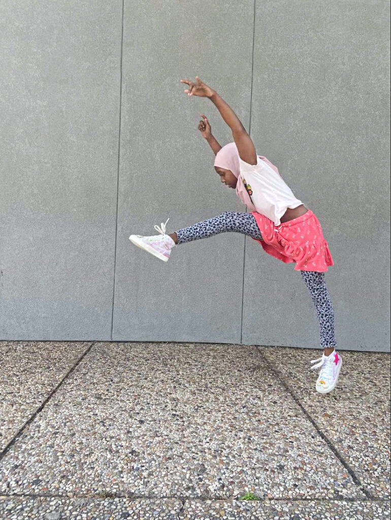 A brown-skinned student wearing a pink headscarf, t-shirt, pink skirt, and leggings does a move with both arms and one leg lifted.