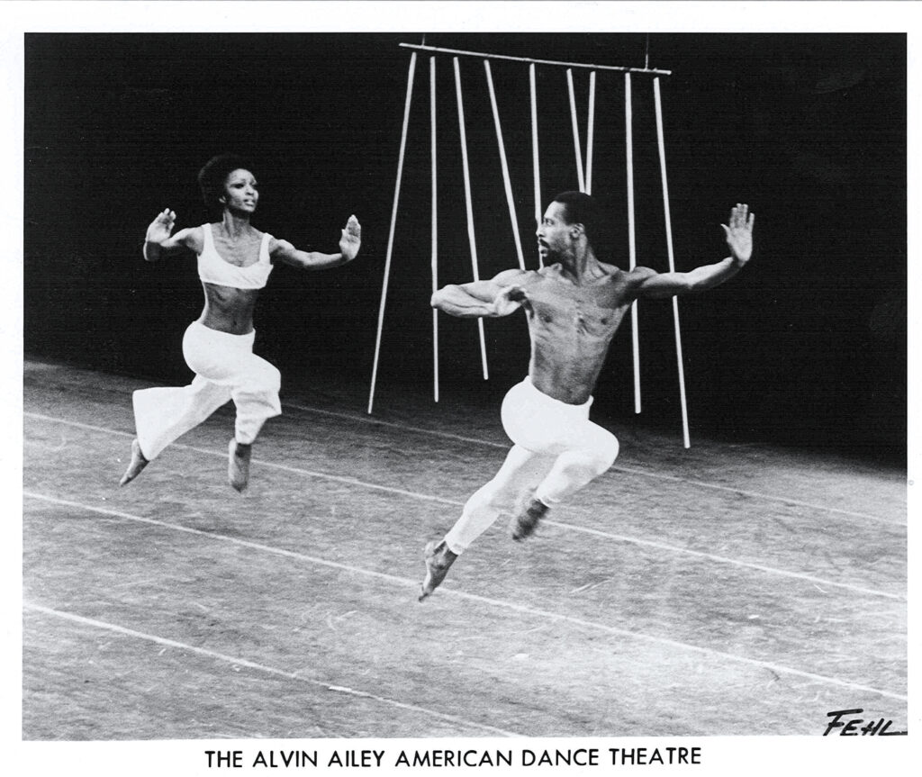 A black and white photo of two Black dancers, a man and a woman, leaping across a stage. He wears white pants and no shirt, and she wears white pants and a white tank top.
