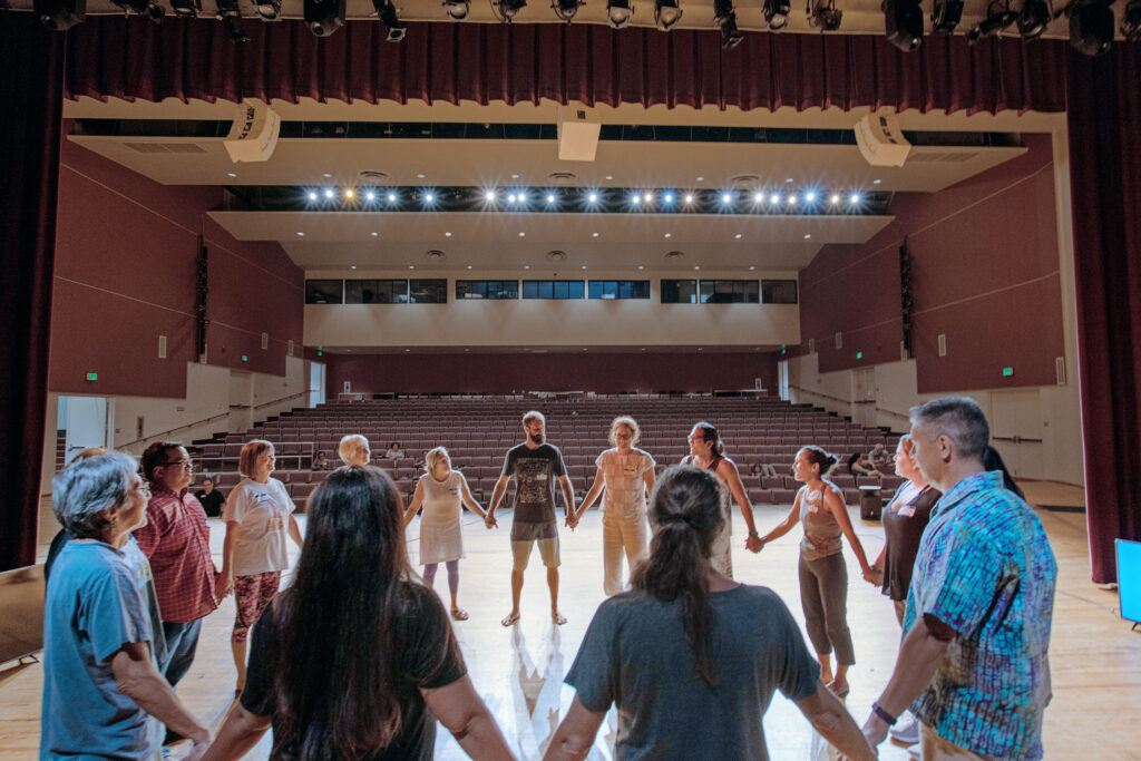 A diverse group of adults holds hands in a large circle onstage in a theater.