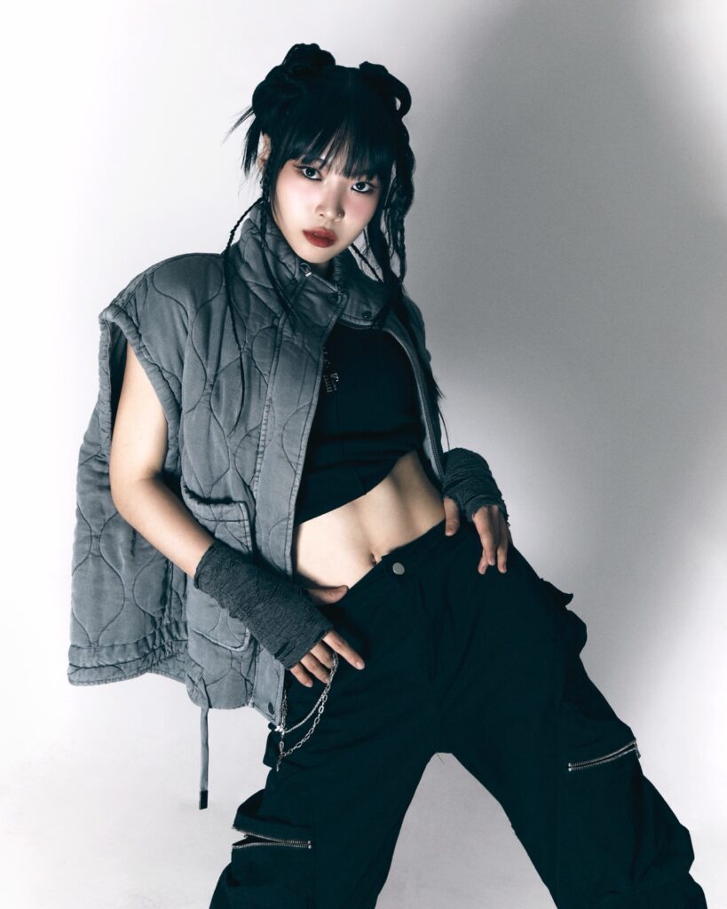 Jo Na-In leans back, hands in her pockets, and juts out one hip as she poses for a portrait. She peers into the camera with her hair up, wearing black pants, a black crop top, a gray vest, and gray arm bands.
