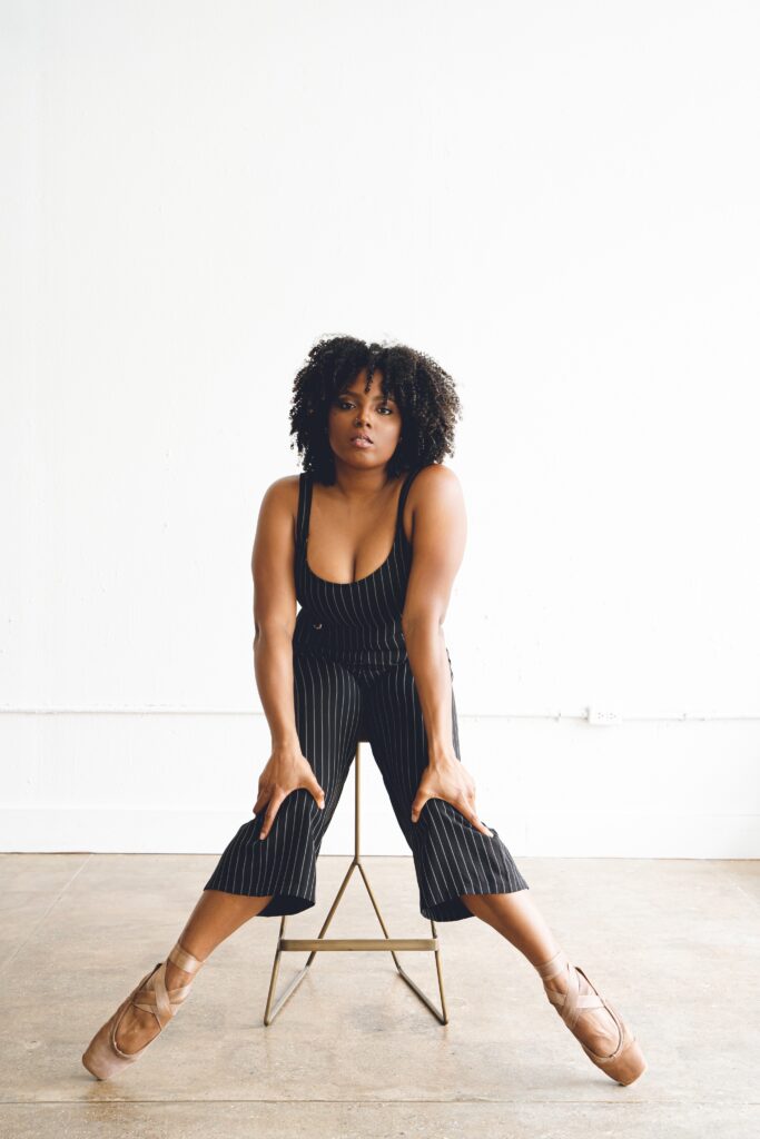Alex Maureen Newkirk, in pointe shoes and a black pantsuit with white stripes, sits on a gold stool with her feet outward, pointed, and knees turned in. She rests her hands on her knees and looks toward the camera with a serious expression. Her natural hair is down.