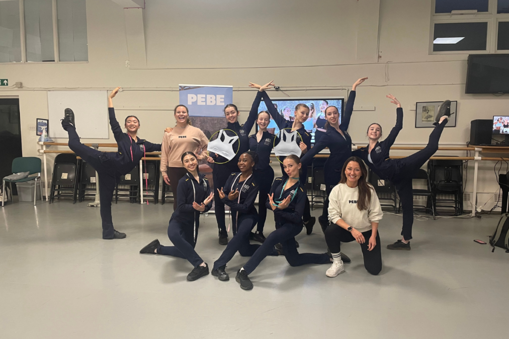 In a ballet studio, two sports bra representatives pose with a group of female ballet students in matching navy athletic sets. Two dancers hold white sports bras, and the two outermost dancers pose in attitude derriere.