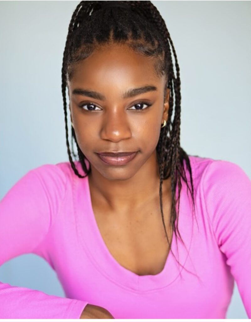 A headshot of Anaya Jones, a Black dancer with long dark braids in a high ponytail. She wears a bright pink long sleeve shirt and looks into the camera with a close-mouthed smile.