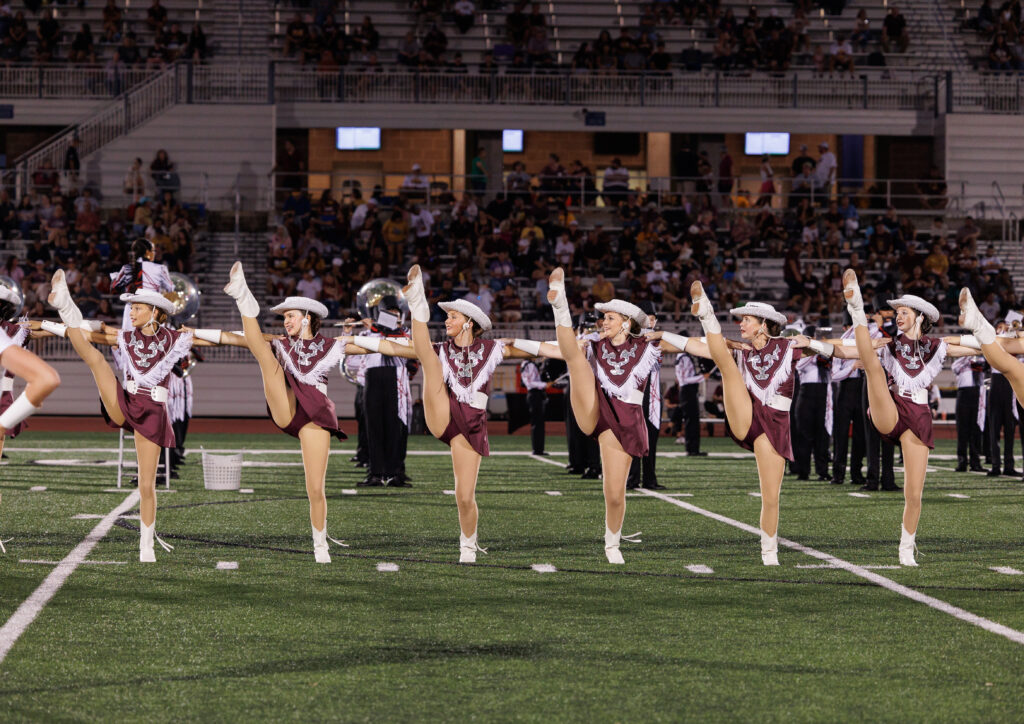 On a football field, members of a drill team form a kick line. They wear maroon dresses with fringe, cowboy hats, and boots.