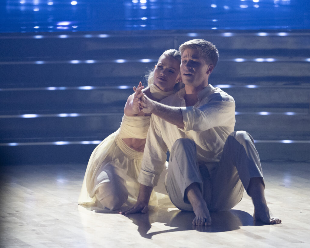 On set at "DWTS," Witney Carson and Robert Irwin sit side-by-side on the floor, knees bent, and hold hands with their arms straight out in front. They have emotional expressions, and they lean their heads against one another's.