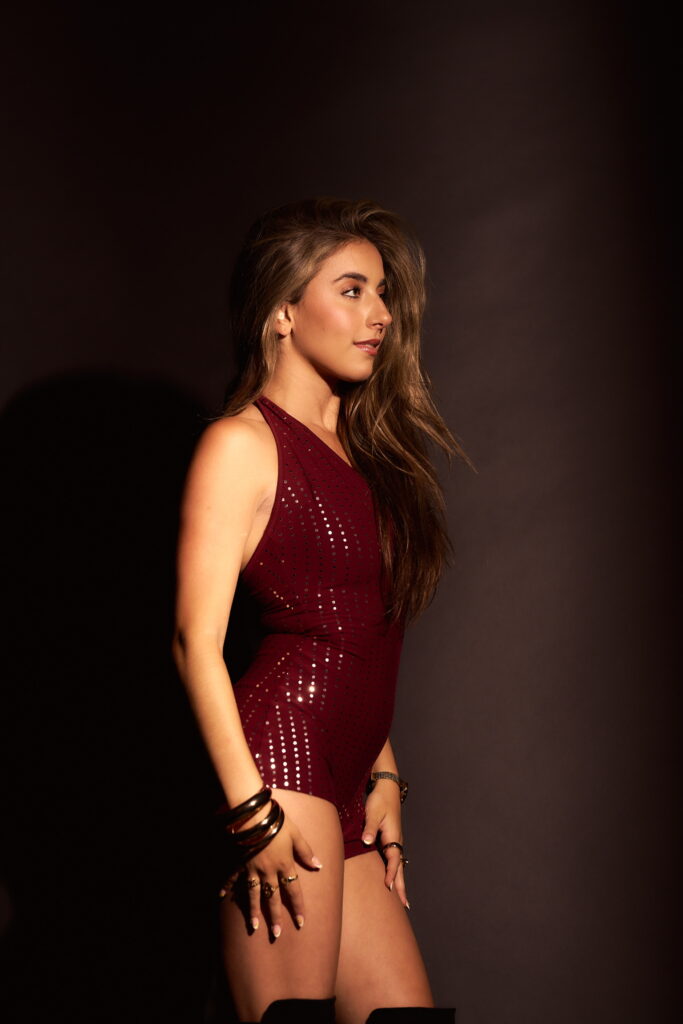In front of a dark brown backdrop, GiaNina Paolantonio stands facing profile, hands resting on the sides of her legs. She wears tall black boots and a short maroon romper that shimmers.
