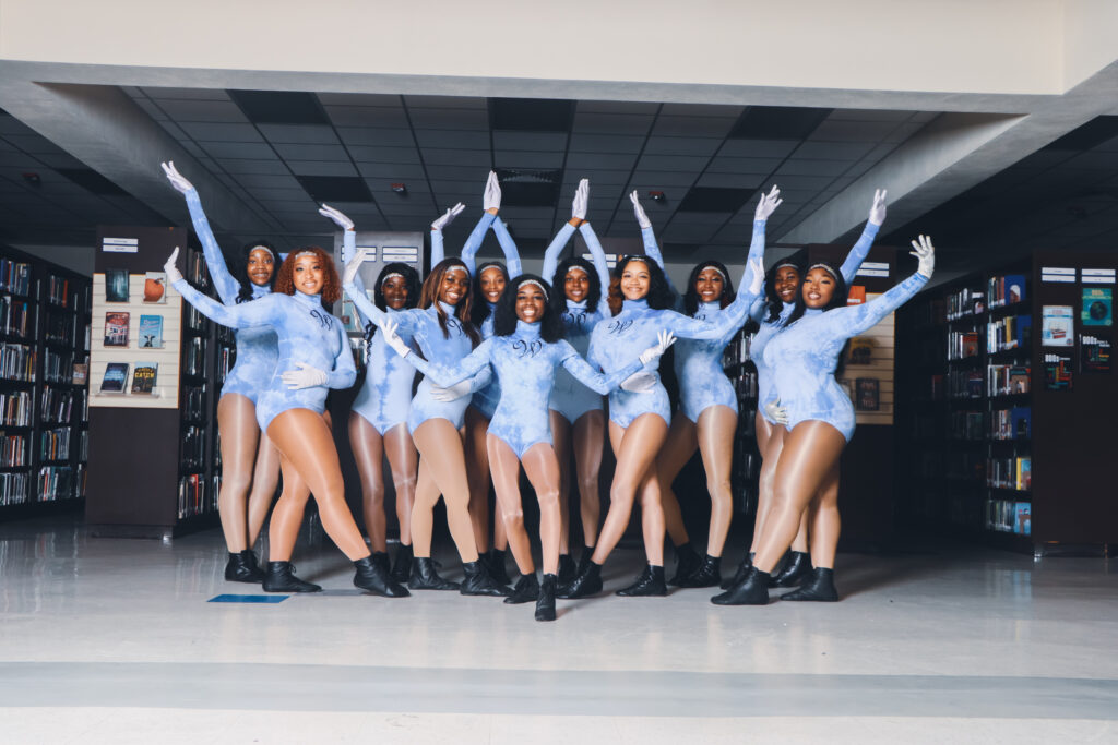 In a school hallway, a majorette team poses for a group photo with their arms lifted. They wear long-sleeved light blue leotards, tan tights, and black shoes.