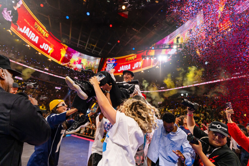 Surrounded by a celebratory crowd and bright marquees, Jaira Joy celebrates at the center of a stadium after winning a street dance battle.