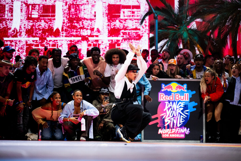 Jaïra Joy dances at a street dance battle, with spectators surrounding her. She crouches low on both legs, arms raised, bent at the elbows, with wrists connected.