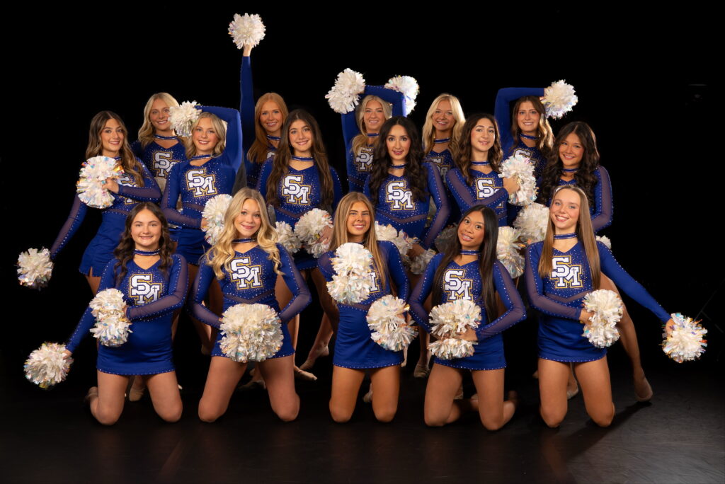 In front of a black backdrop, members of a song team pose together holding white poms and wearing royal blue uniforms.