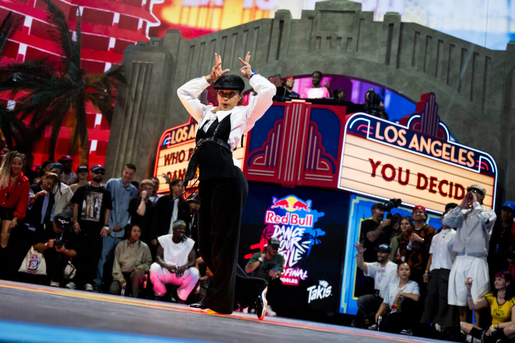 Jaïra Joy dances at a street dance battle, surrounded by bright marquees and spectators. She stands with style, arms raised with elbows bent, wrists touching either side of her head as her fingers point up.