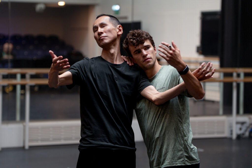 In a ballet studio with their backs to the mirror, Takashi Kikuchi and Leo Dixon dance together side-by-side, Dixon resting an arm and his head on Kikuchi’s left shoulder. Kikuchi, eyes closed, lifts his arms up, bent at the elbow, and Dixon raises his other arm, looking at his palm intently.