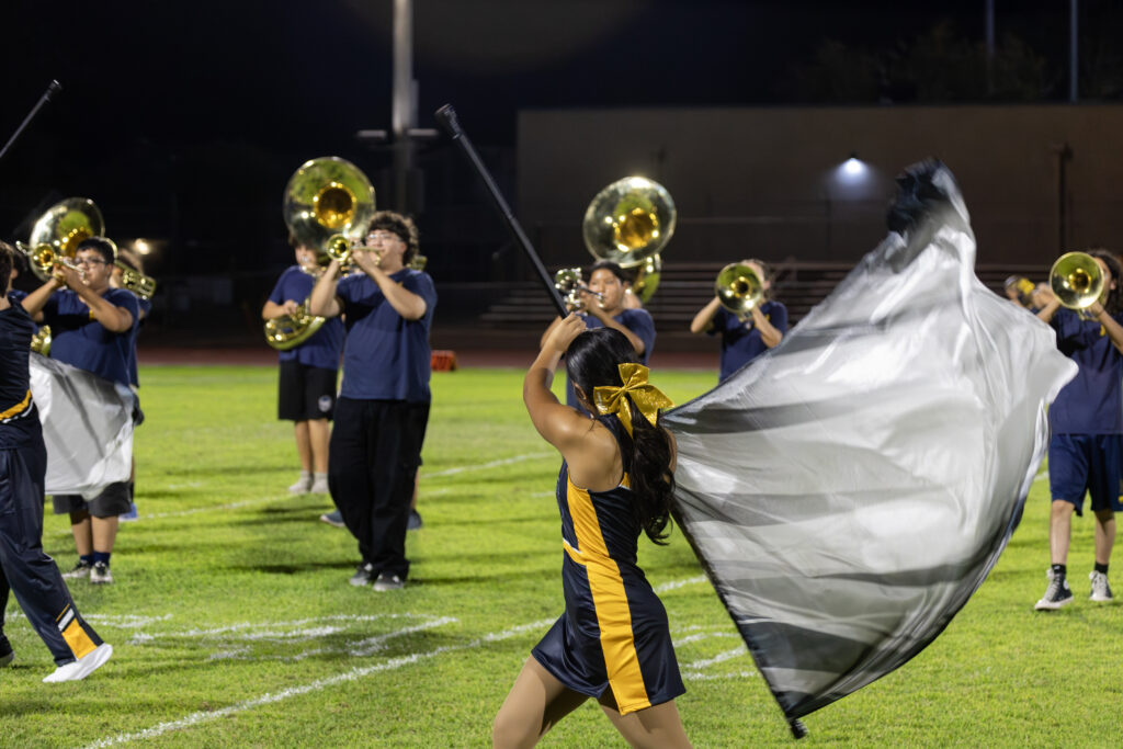 On a football field, band members play as a member of the color guard walks forward, spinning a large silver flag.