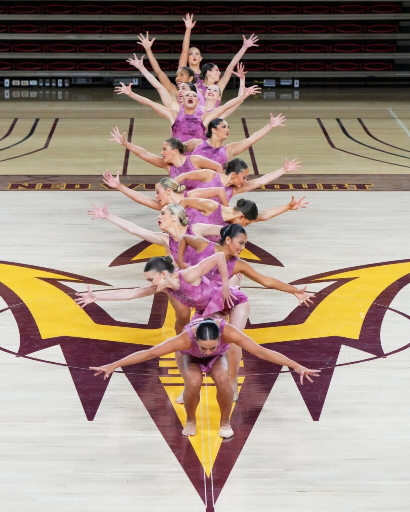 On the ASU basketball court, the dance team forms a vertical line as they extend their limbs in opposite directions. They wear dark pink costumes with short skirts
