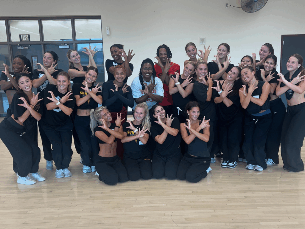 In a dance studio, the ASU dance team members pose for a group photo with Toya Renee Ambrose. They all make the ASU sun devils sign with their hands as they smile.