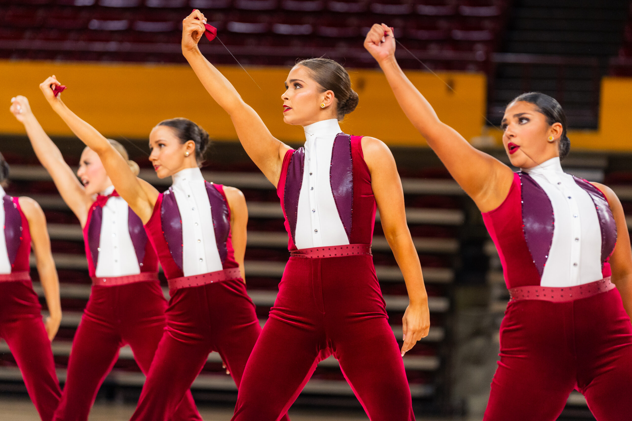 A close-up of members of ASU's dance team, who strike the same pose with legs wide and one arm extended upward, pulling a rose from their lapel. Their costumes are red velvet pants and vests with a white shirt underneath.